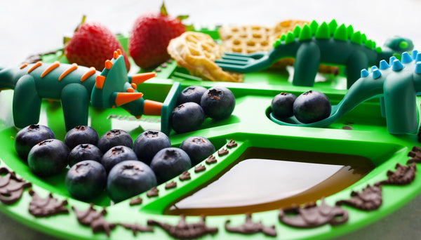 Dinosaur green plate with healthy yummy food on dinosaur section plate with Triceratops pusher pushing healthy food onto Ankylosaurus spoon and Stegosaurus fork picking up healthy eating habits